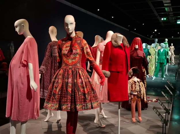 Ausstellung von Modedesigns auf Schaufensterpuppen, präsentiert in verschiedenen Farben und Stilen, darunter ein rotes Kleid mit einem gemusterten Unterrock.